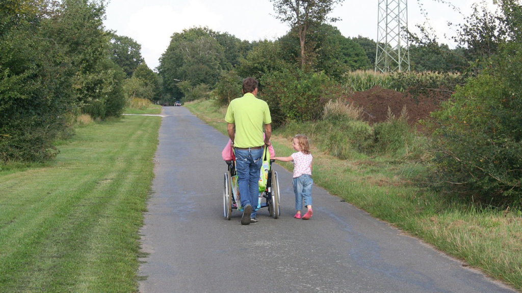 Eine Familie geht im Grünen spazieren. Der Vater schiebt den Rollstuhl, in dem die Mutter sitzt. Die Tochter geht neben dem Rollstuhl.